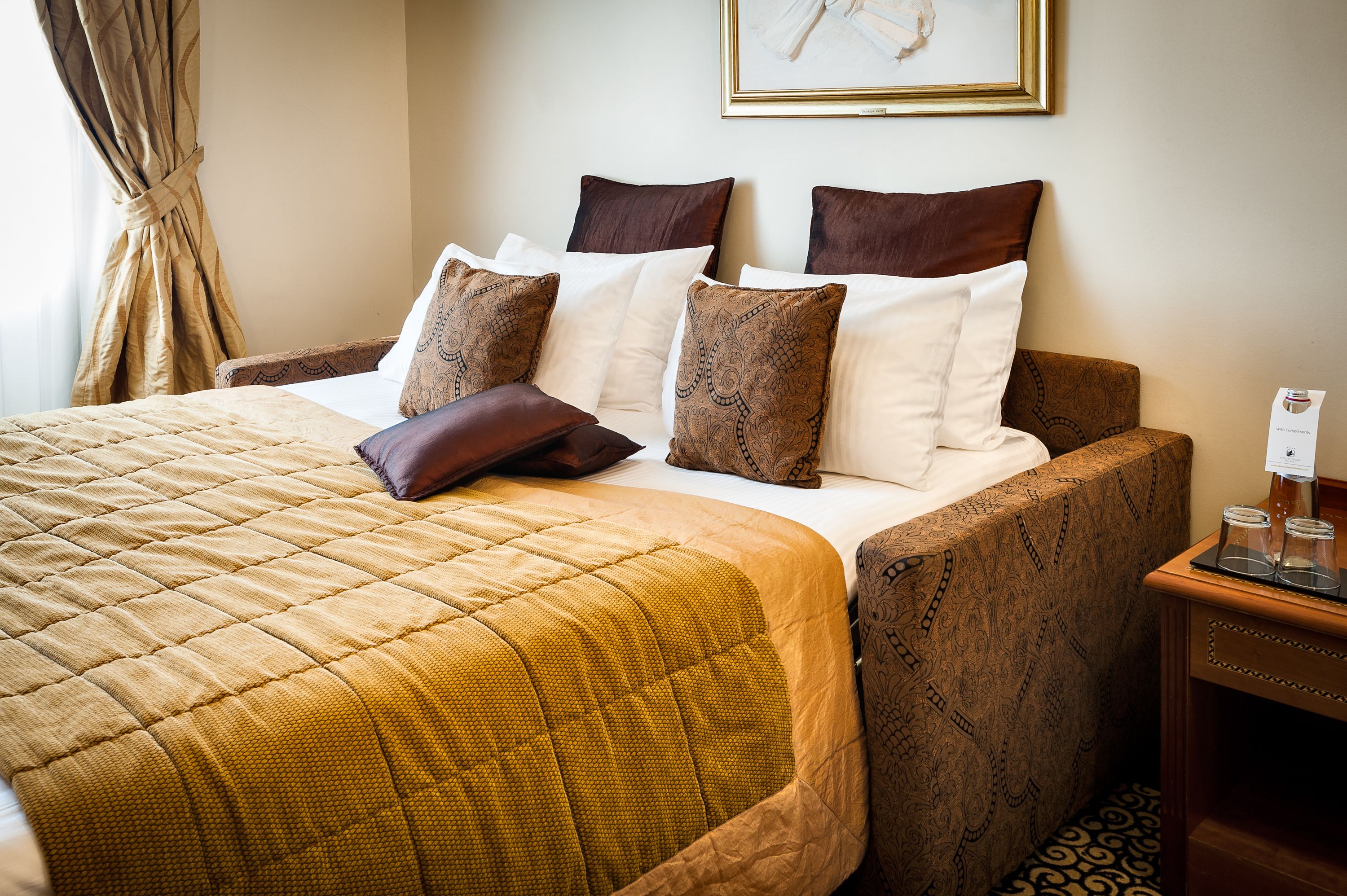 Un letto rifatto con cura, con copripiumino e cuscini decorati, una testiera in legno e un comodino con un bicchiere e un piccolo contenitore.