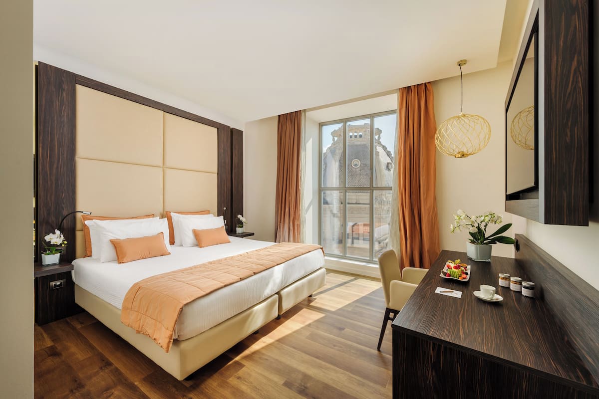 Habitación de hotel moderna con una cama grande, una mesa de comedor y vistas a un edificio histórico a través de la ventana.