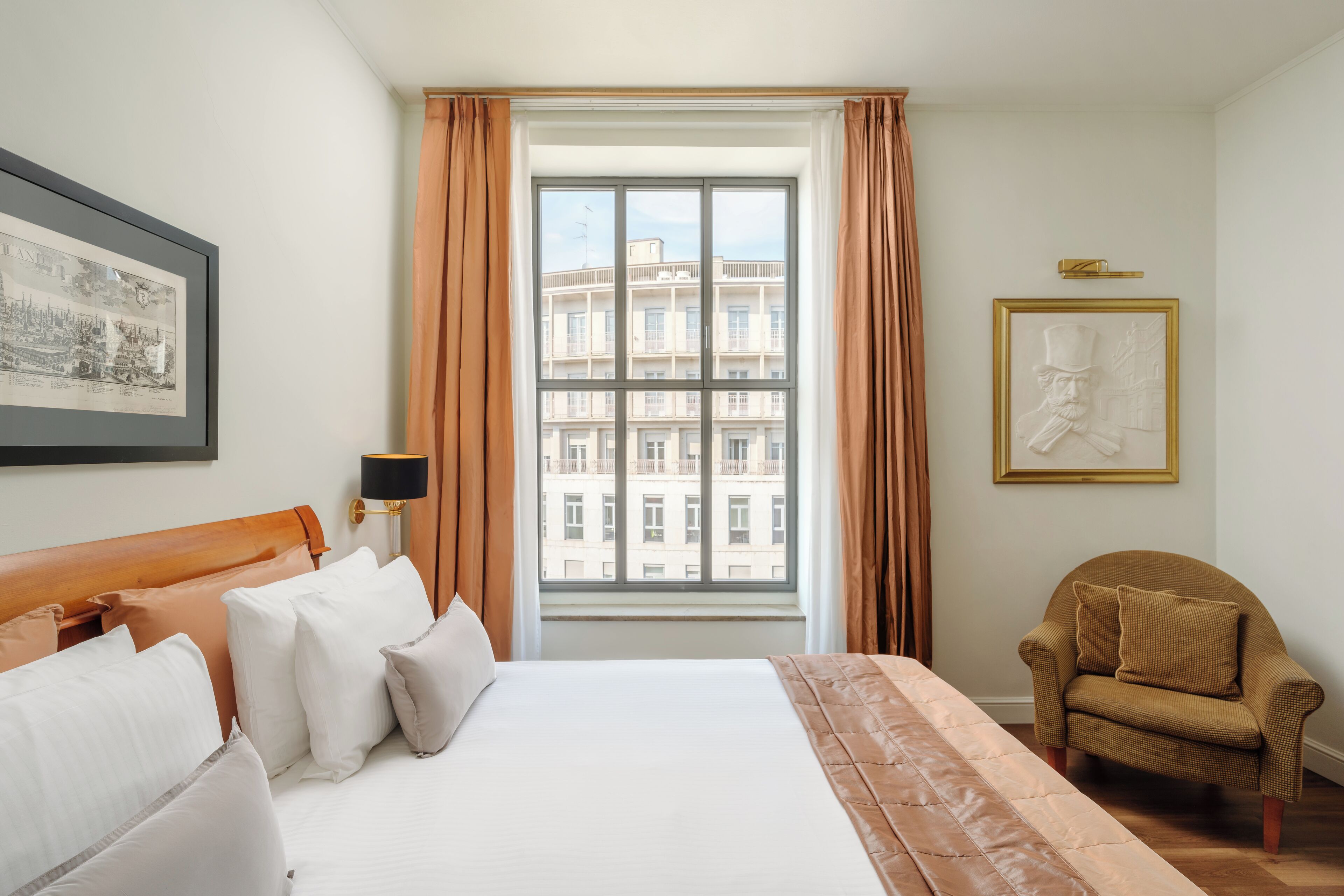 A hotel room with a large bed, a chair, a window with curtains, and framed artwork on the walls.