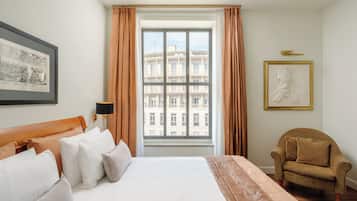 A hotel room with a large bed, a chair, a window with curtains, and framed artwork on the walls.