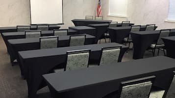 A conference room with multiple tables and chairs, a projection screen, an American flag, and a window with blinds.