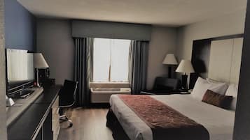 A hotel room with a bed, desk, chair, two lamps, a TV, and an air conditioning unit.