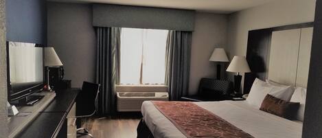A hotel room with a bed, desk, chair, two lamps, a TV, and an air conditioning unit.