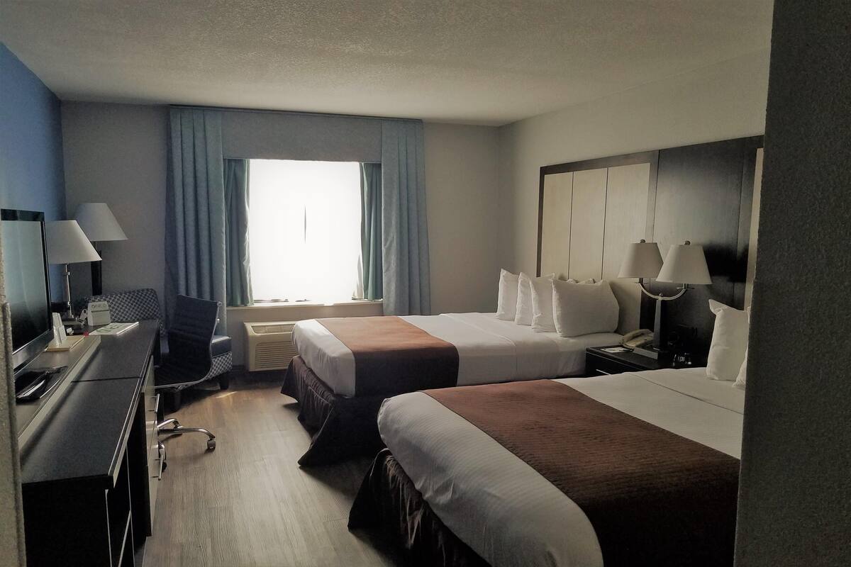 Habitación de hotel con dos camas, un escritorio con computadora, una silla, ventana con cortinas y un espejo grande.