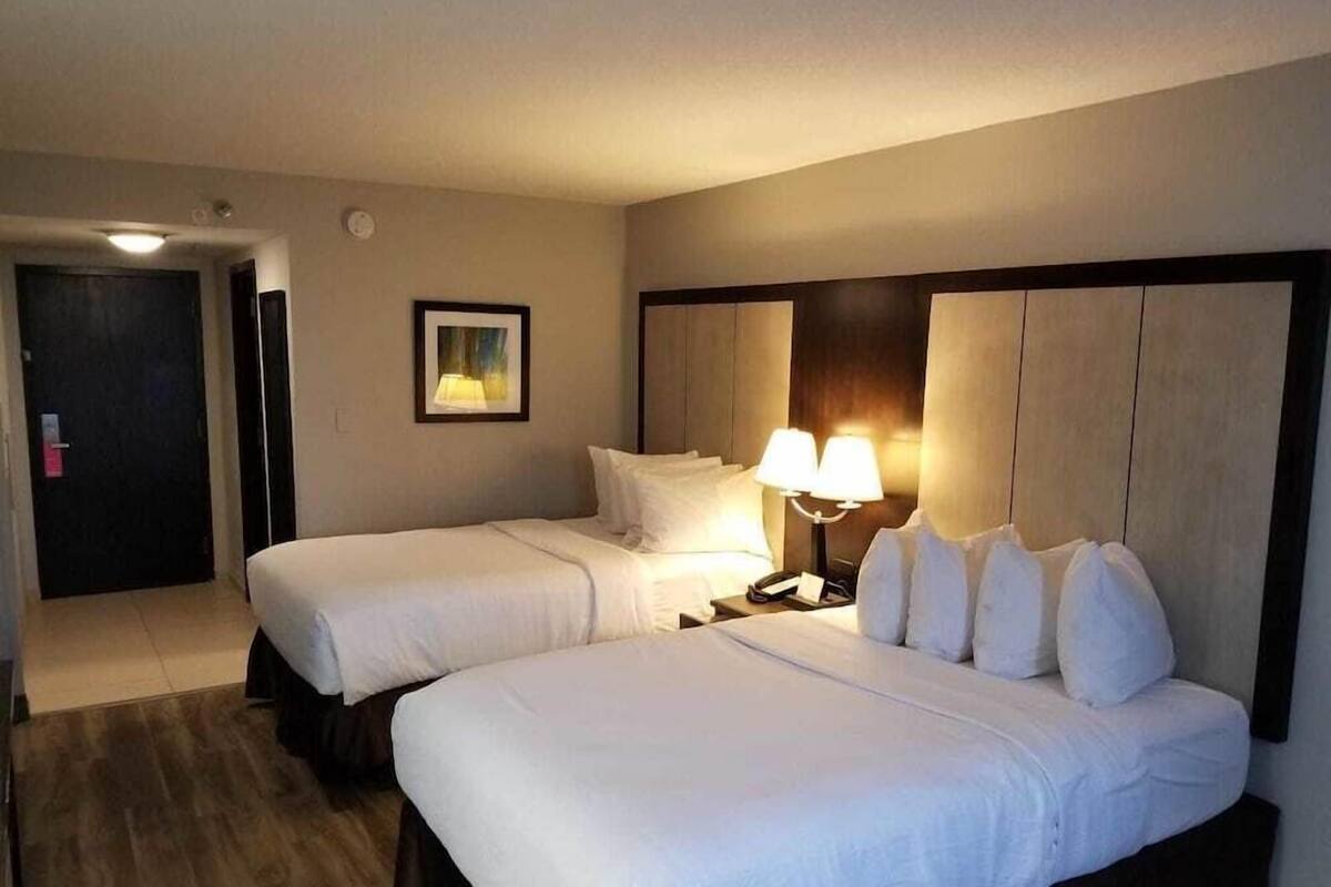 Habitación de hotel con dos camas, una mesita de noche con lámpara, un cuadro en la pared y una puerta al fondo.