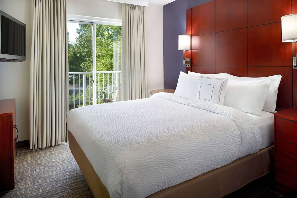 Hotelkamer met een groot bed, een aan de muur bevestigde televisie, een raam met gordijnen en uitzicht op groen.