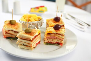 Ein Teller mit gestapelten Sandwiches, Oliven und Kirschen, eine Schüssel Pommes und ein Glas Milch auf einem weißen Tisch.