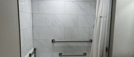 Chuveiro com azulejos de mármore branco, porta de vidro e barra de apoio metálica.