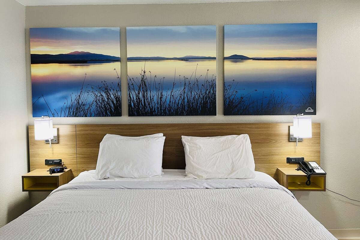 A hotel room with a large bed, two bedside tables, a nightstand, a wall-mounted phone, and a painting of a landscape.