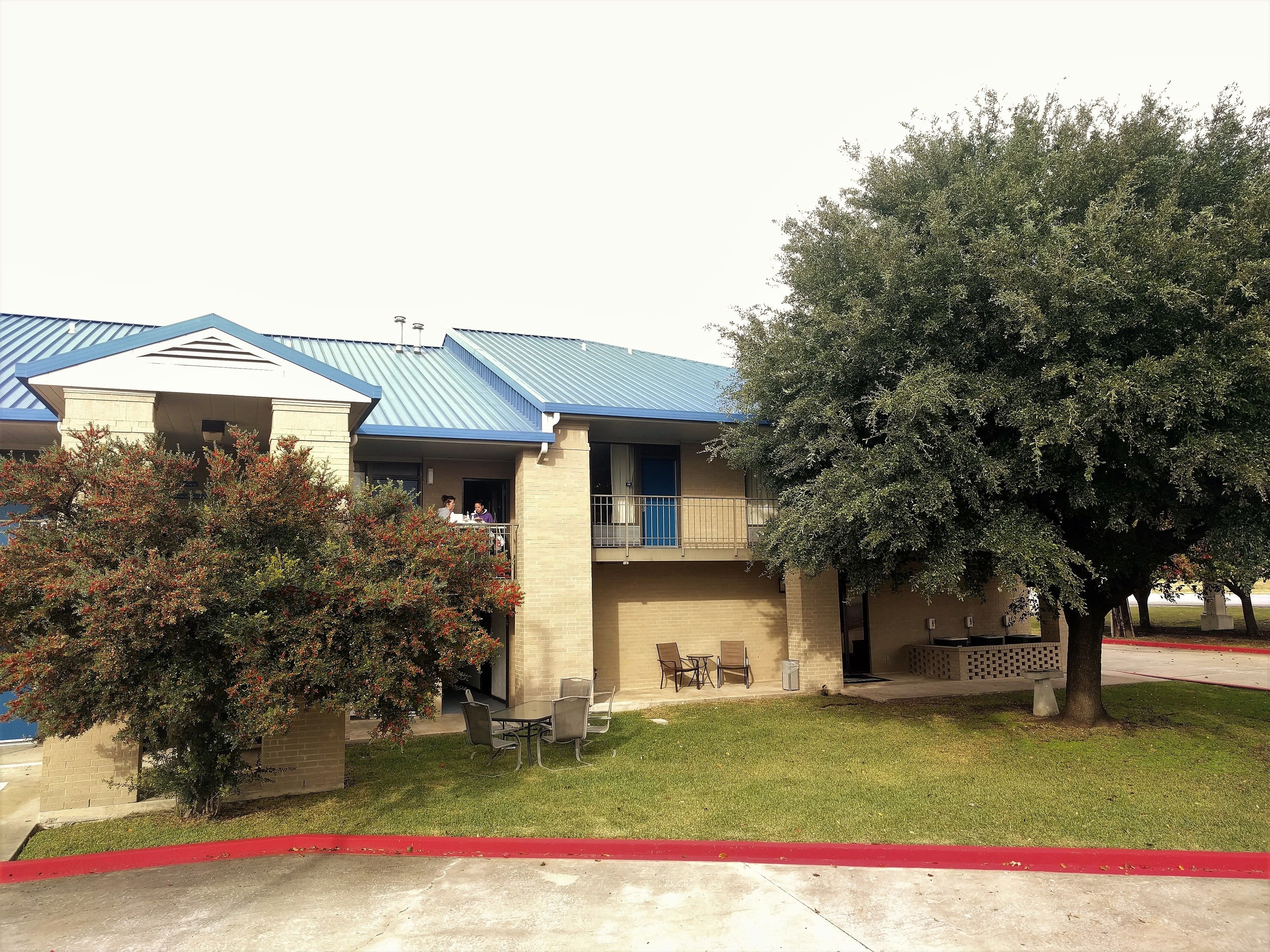 A two-story building with a blue roof, a balcony, and a small outdoor seating area with tables and chairs.