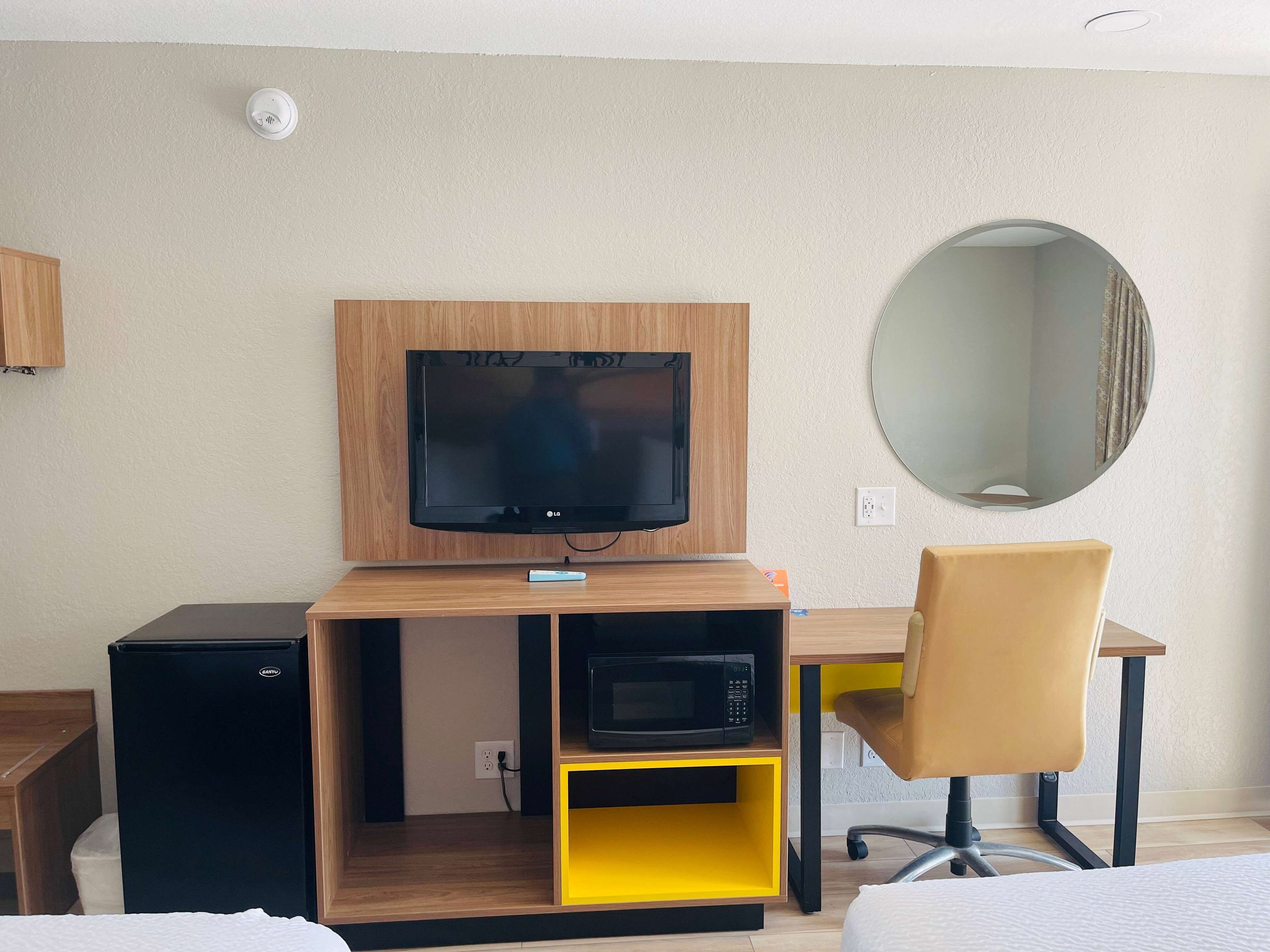 A hotel room with a flat-screen TV mounted on a wooden cabinet, a microwave, a small refrigerator, a yellow storage shelf, a round mirror, and a brown chair.