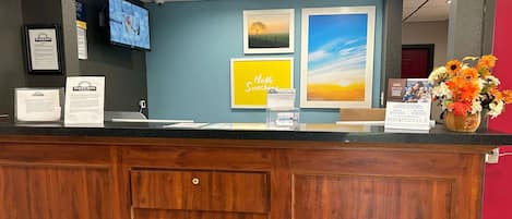 A hotel reception desk with a wooden counter, framed informational posters, a digital screen, and a vase of flowers.