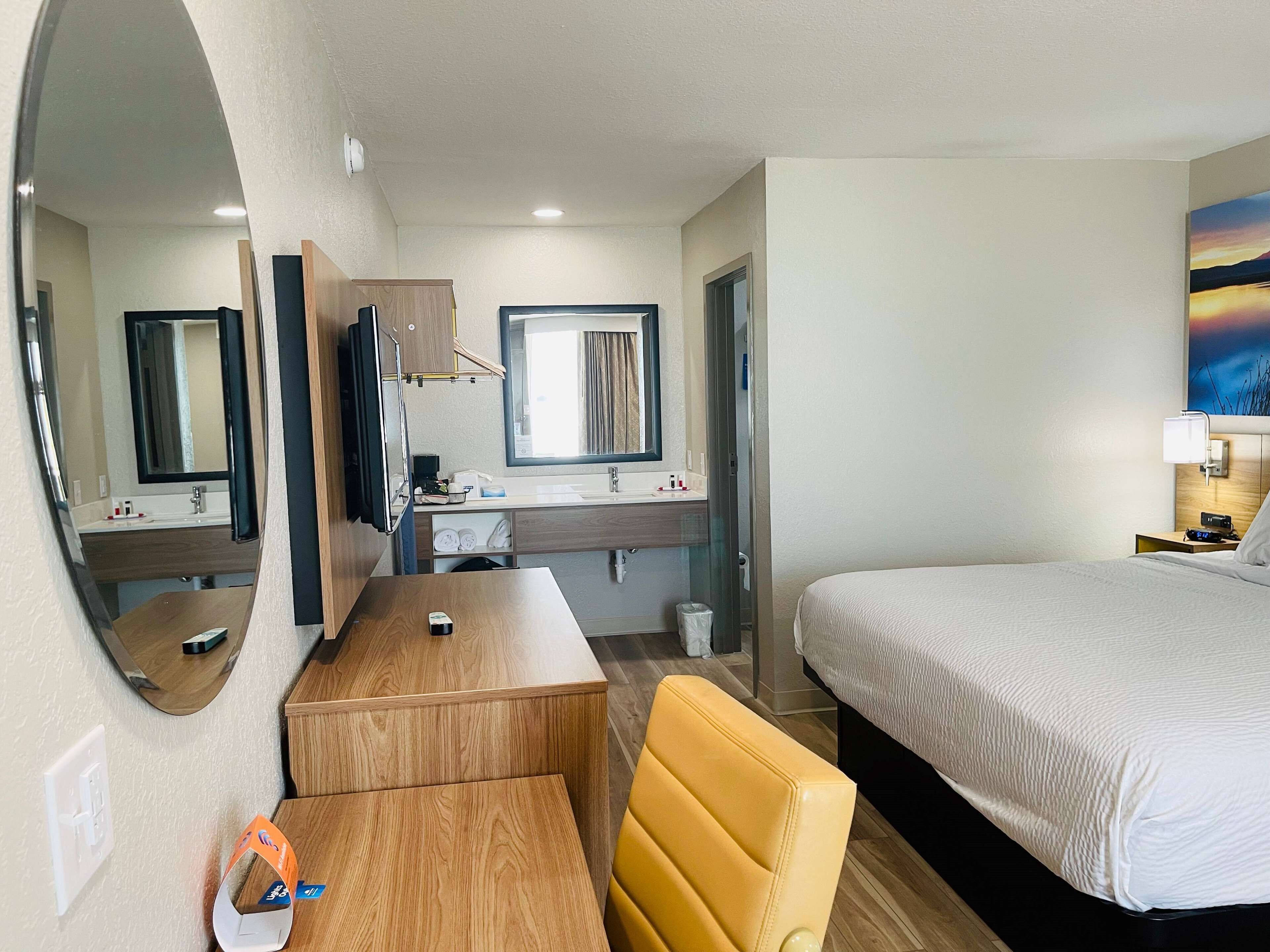 Une chambre d’hôtel avec un lit, un bureau, une chaise, une télévision, un miroir, un lavabo et un tableau au mur.