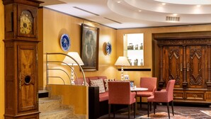 A hotel lobby with a wooden staircase, red upholstered seating, a dining area with red chairs, and a large wooden cabinet with decorative carvings.