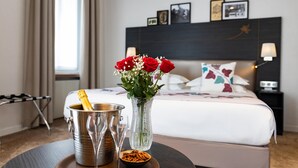 A hotel room with a bed, bedside table, lamp, and a table with a vase of flowers, a champagne flute, and a bowl of snacks.