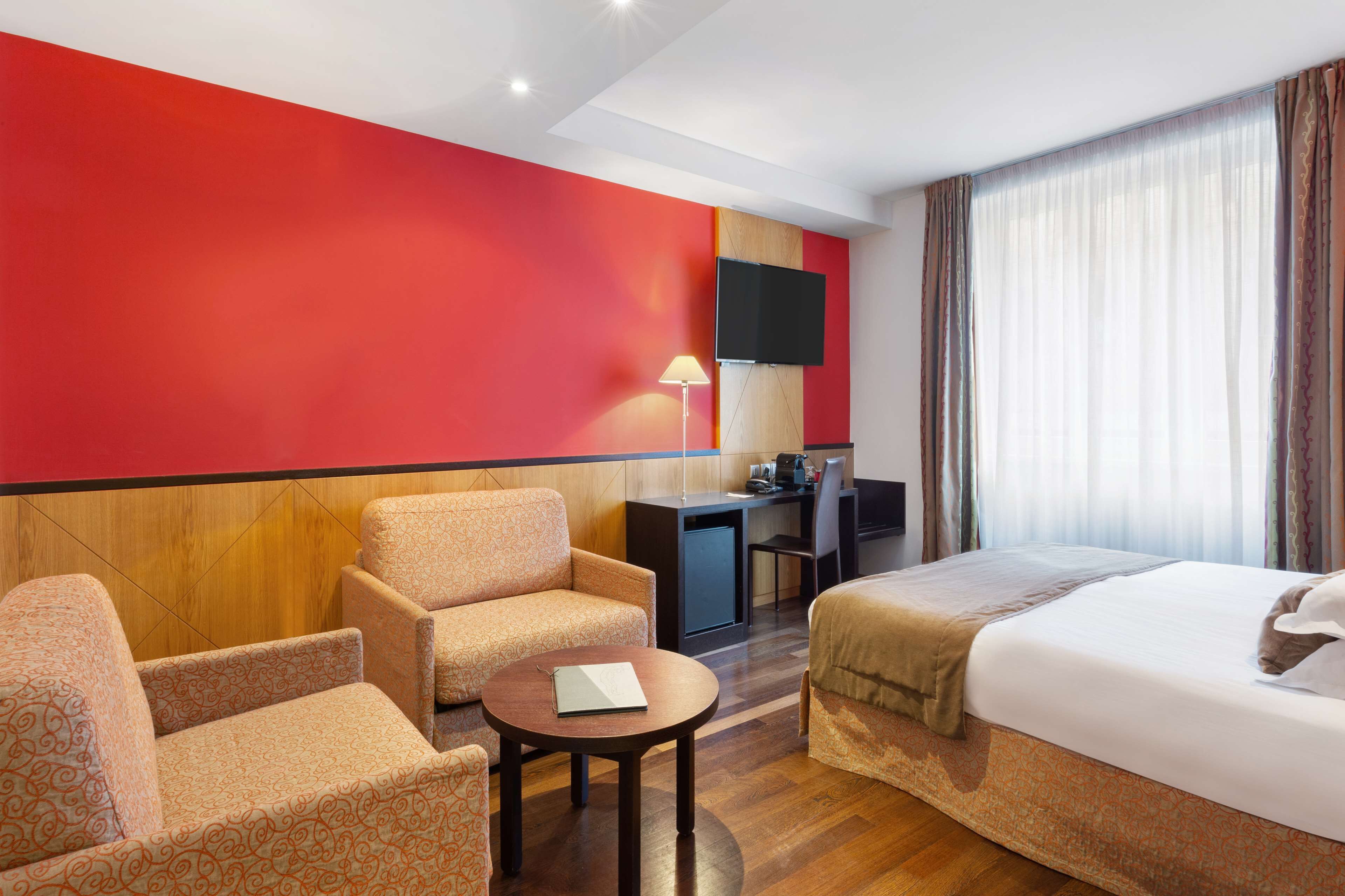 Habitación de hotel con pared de acento roja, una cama, dos sillones, un escritorio con computadora, un televisor montado en la pared y una ventana con cortinas.