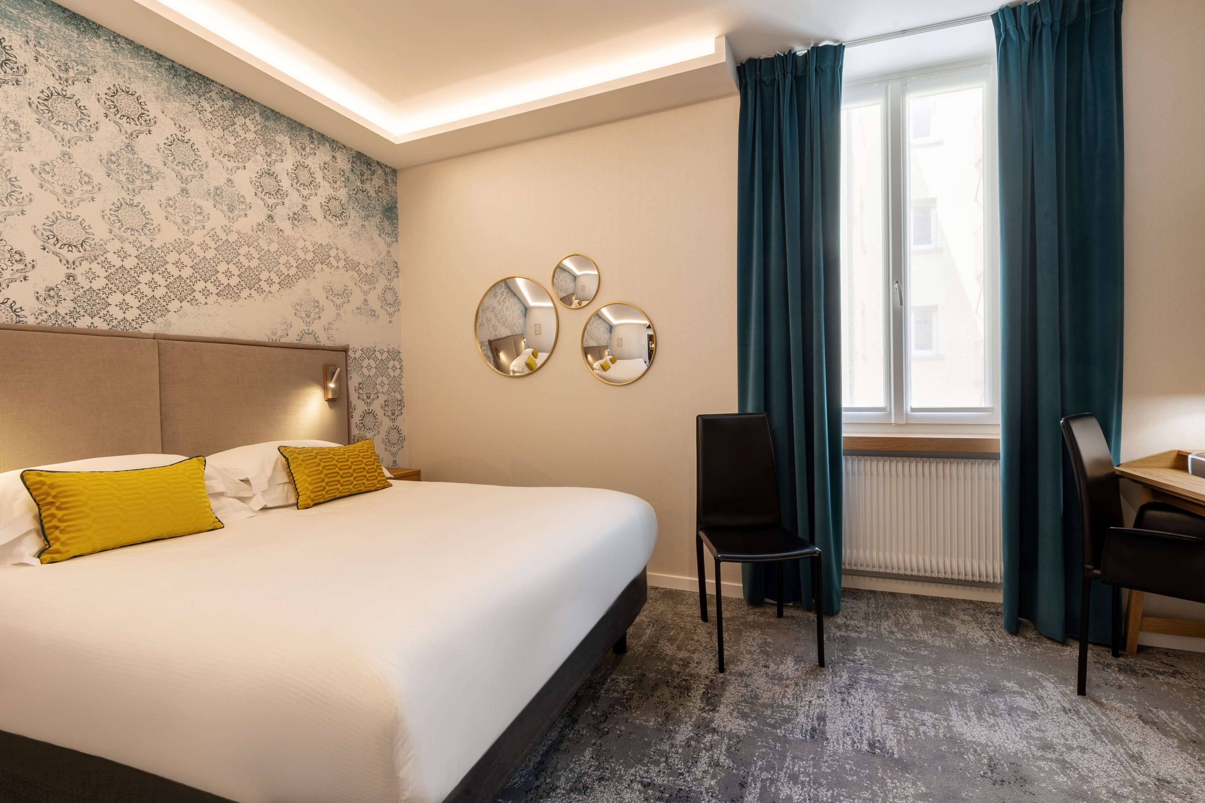 Une chambre d’hôtel avec un lit, deux chaises, un bureau et une fenêtre avec des rideaux.