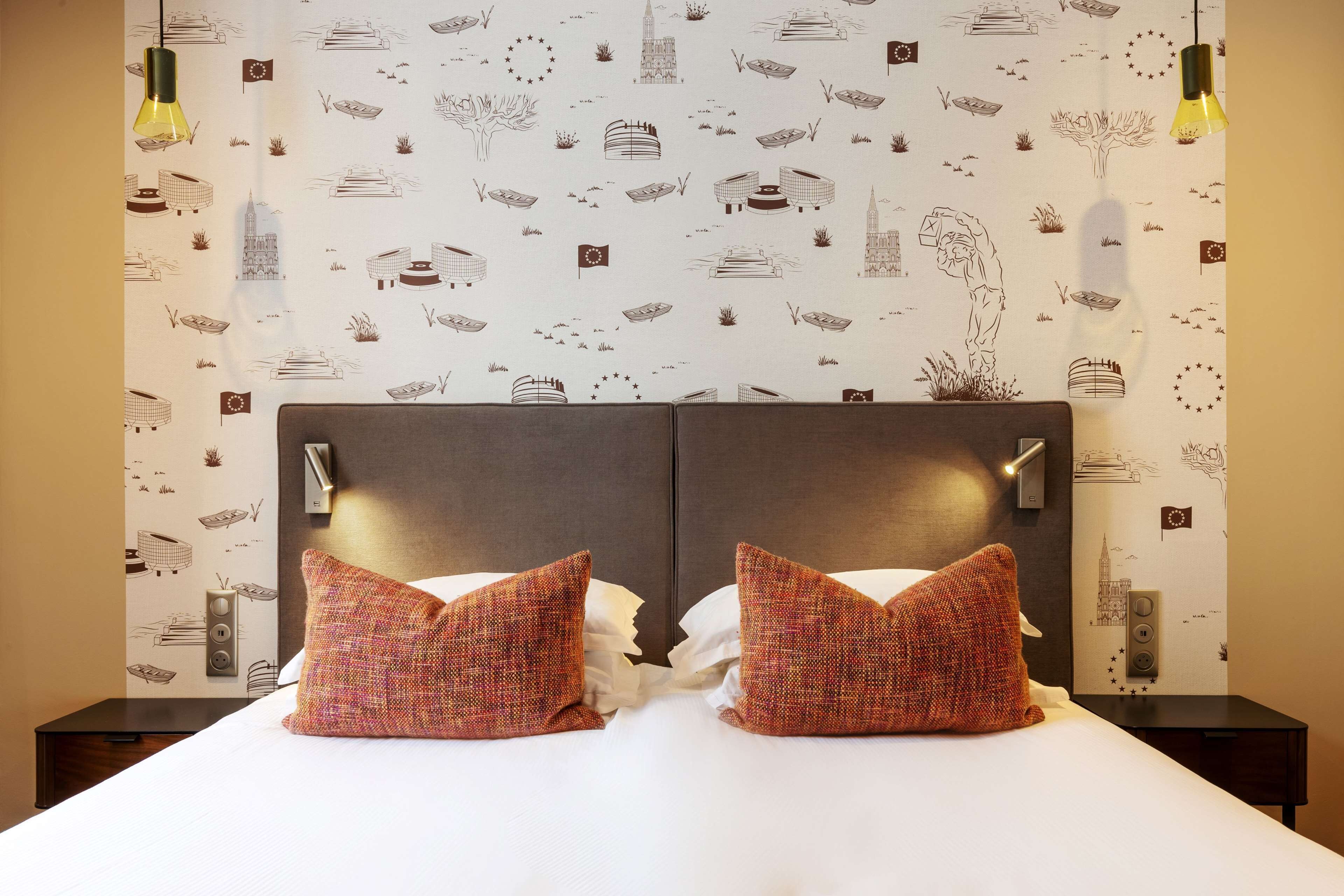 A hotel room with a large bed, two decorative pillows, a nightstand, a wall-mounted light, and a patterned wallpaper featuring architectural drawings.