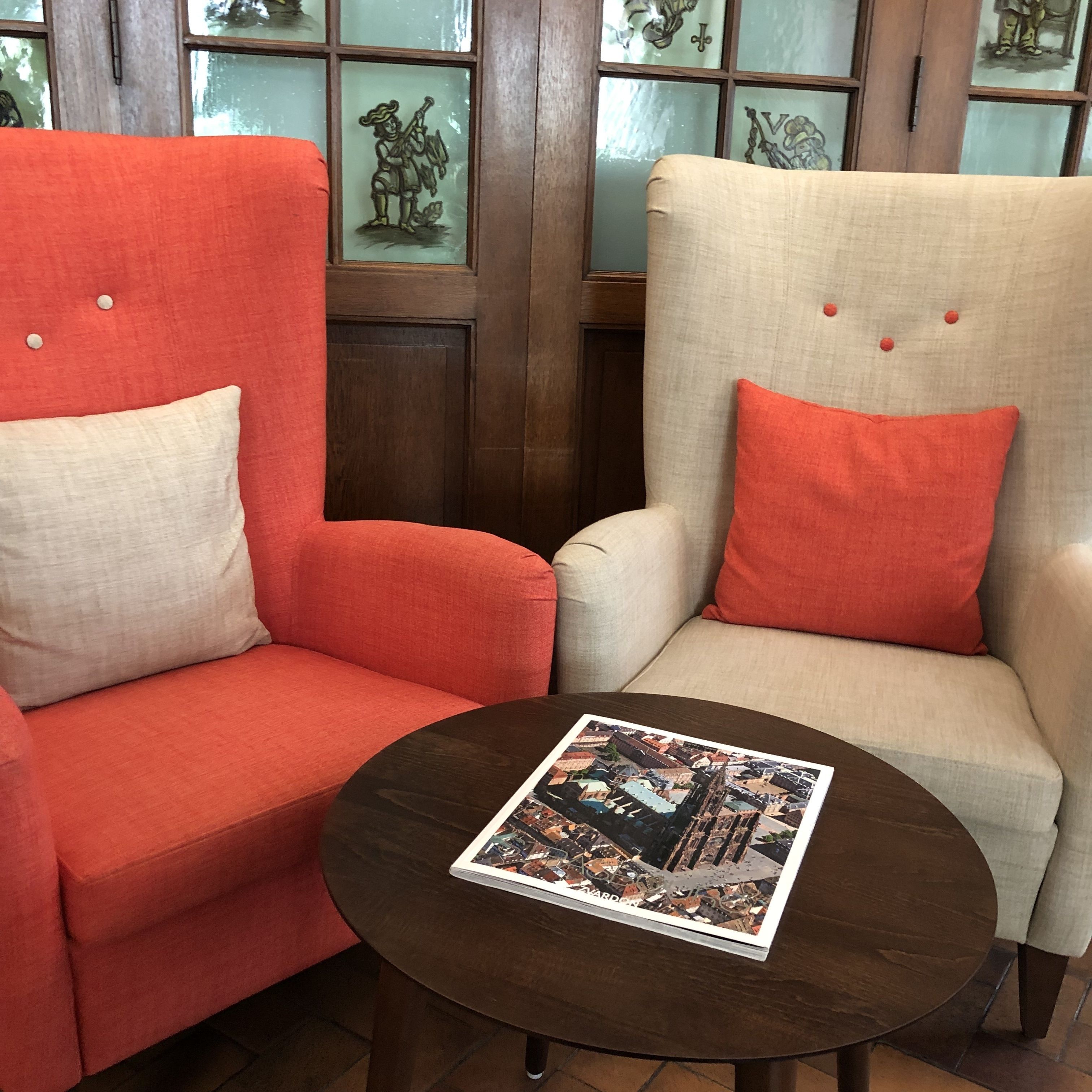 Une pièce comprenant deux fauteuils, l’un rouge et l’autre beige, agrémentés de coussins orange assortis. Une table ronde en bois, sur laquelle repose un magazine, se trouve devant les fauteuils.