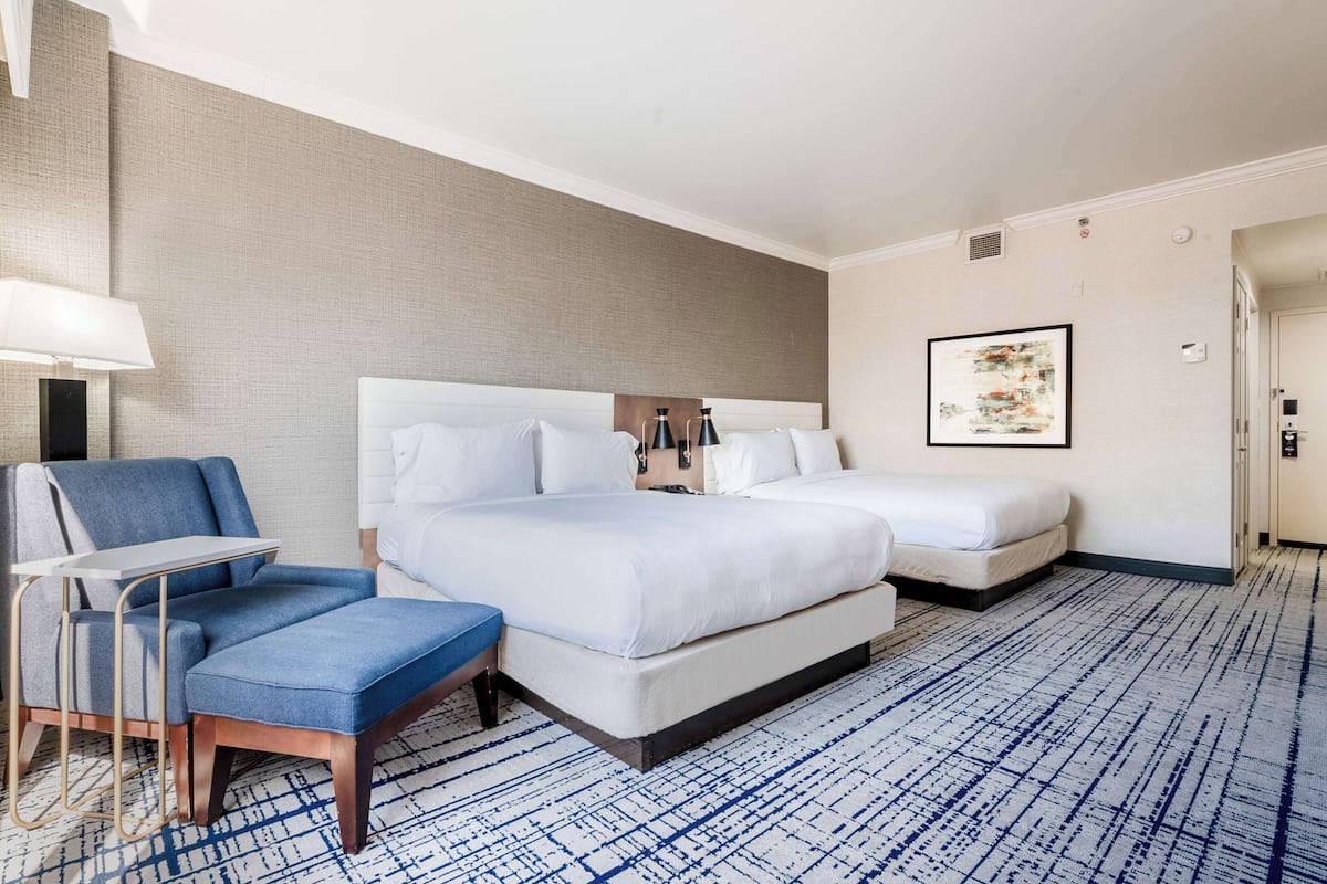 A hotel room with a large bed, a smaller bed, a desk with a chair, and a blue ottoman.