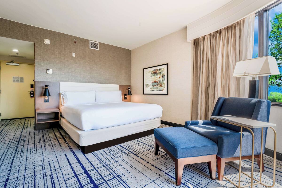 A hotel room with a bed, a desk, a chair, a lamp, and a window with curtains.