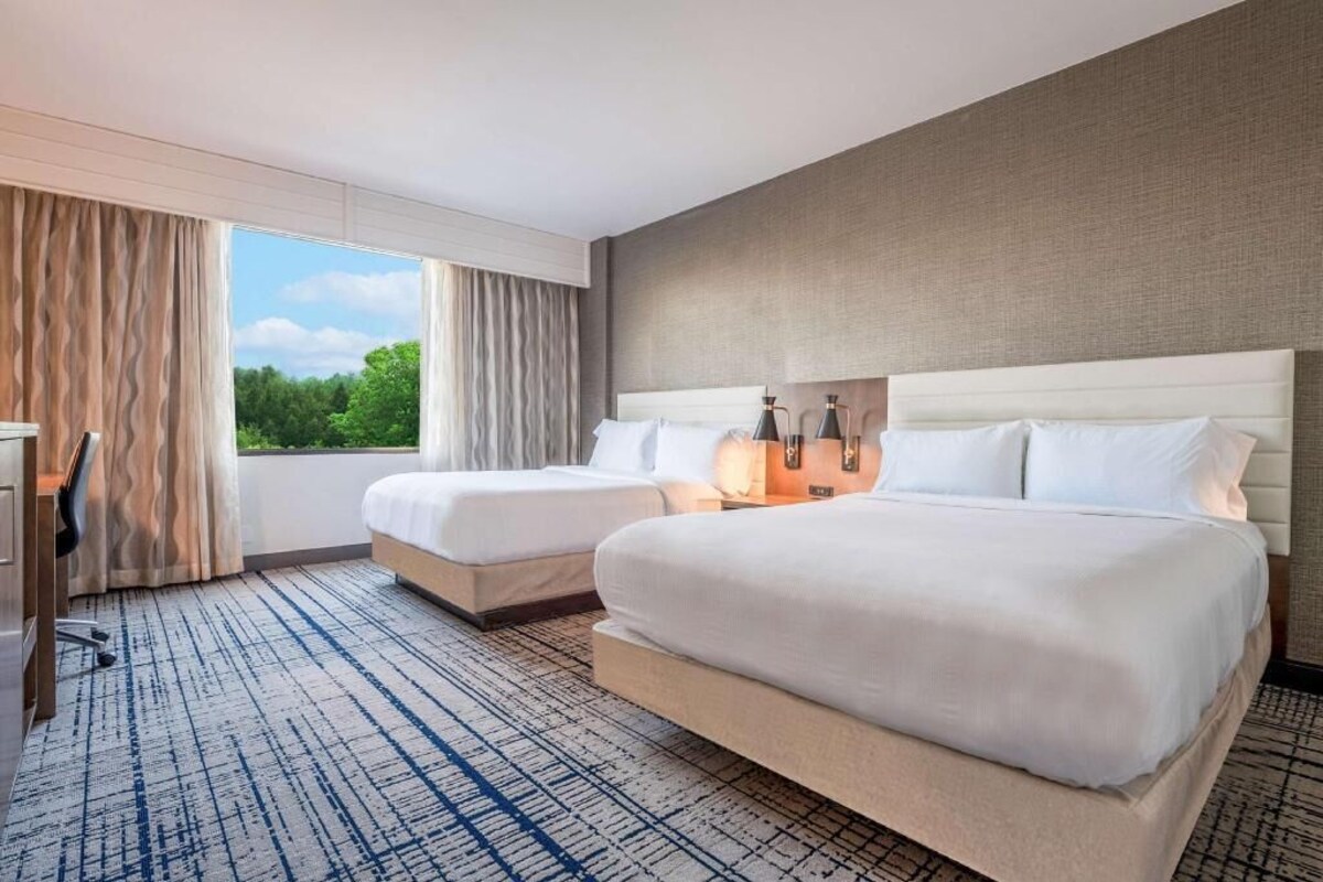 A hotel room with two beds, a desk, and a view of greenery.