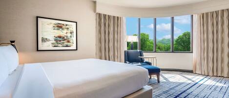 A hotel room with a large bed, a desk, a chair, and a window with a view of greenery.