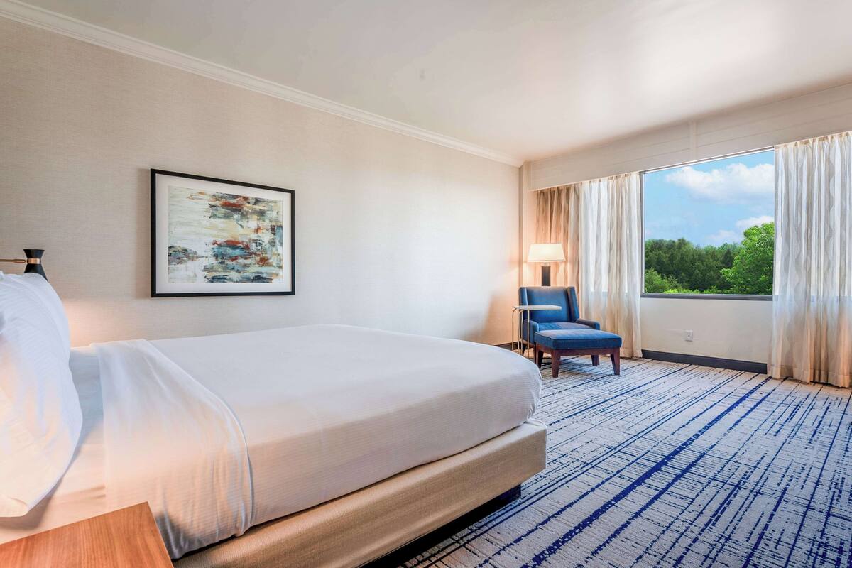 A hotel room with a large bed, a chair, a window with curtains, and a painting on the wall.