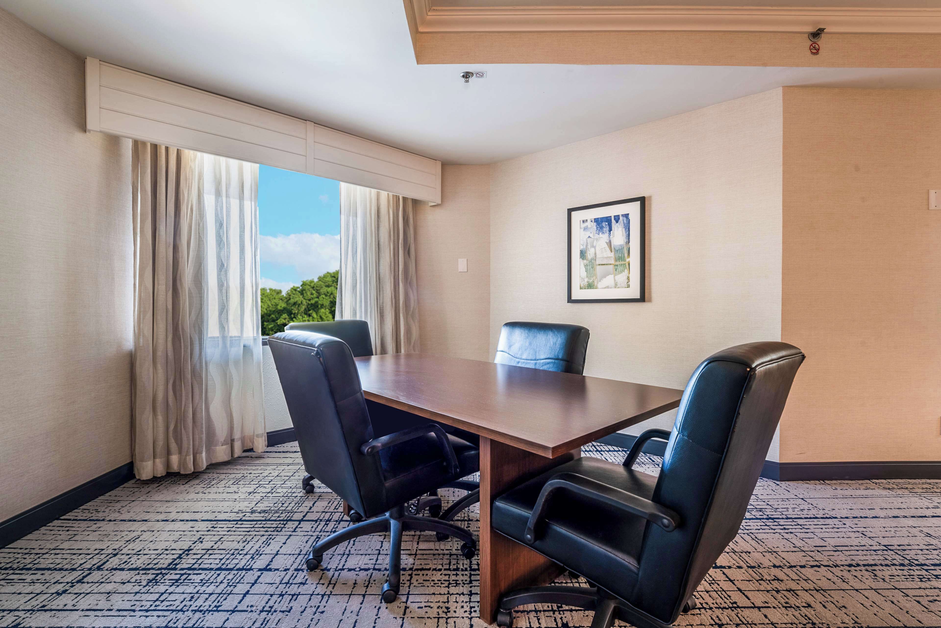 A conference room with a wooden table, four black chairs, a large window with curtains, and a framed picture on the wall.