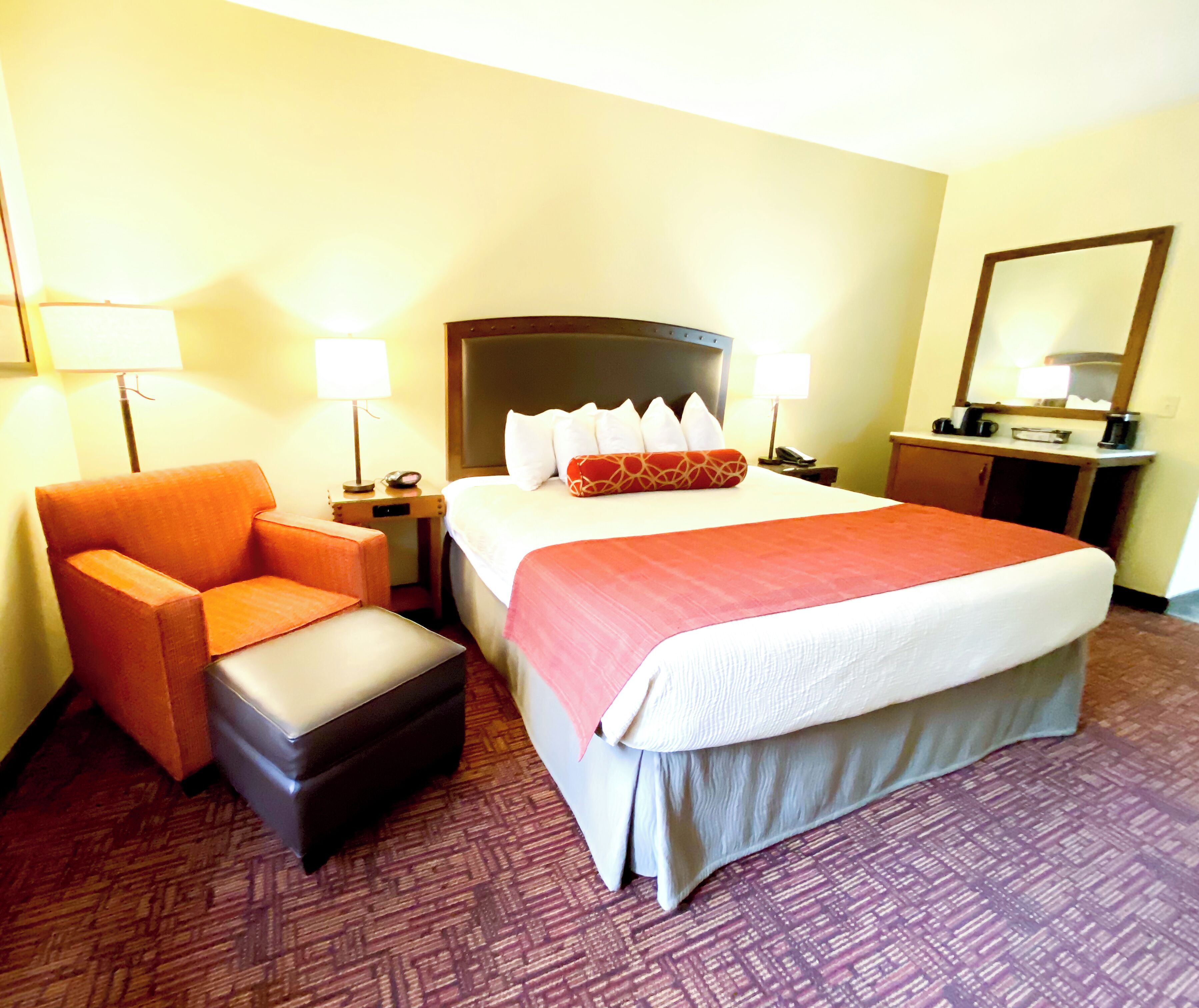 A hotel room with a bed, a chair, a nightstand, a lamp, a desk, and a mirror.