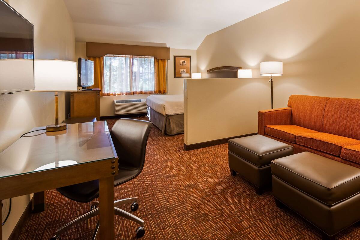 A hotel room with a sofa, ottomans, a desk with a chair, a TV, and a window with curtains.