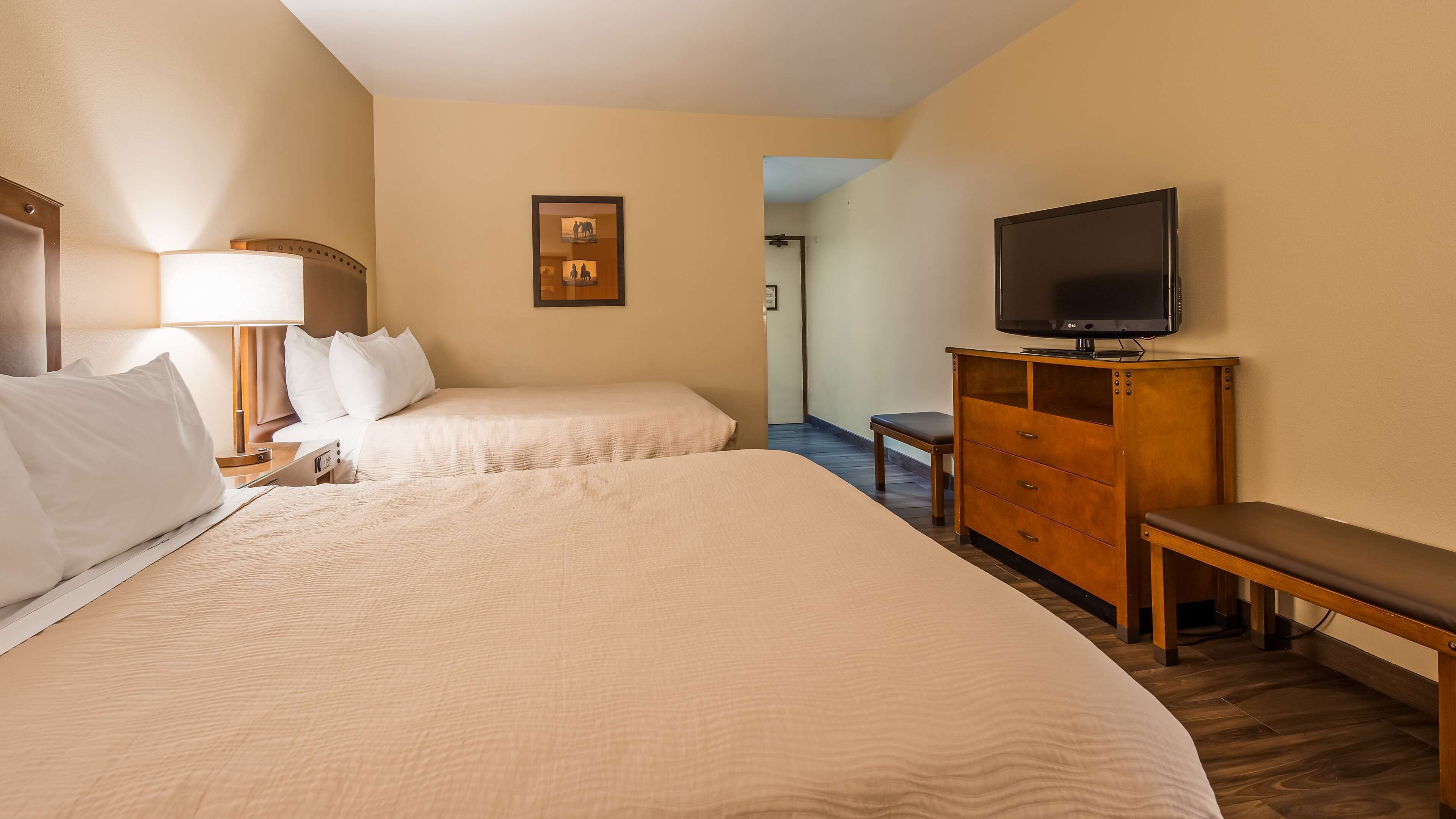 A hotel room with two beds, a TV on a wooden cabinet, a bench, and a desk with a lamp.