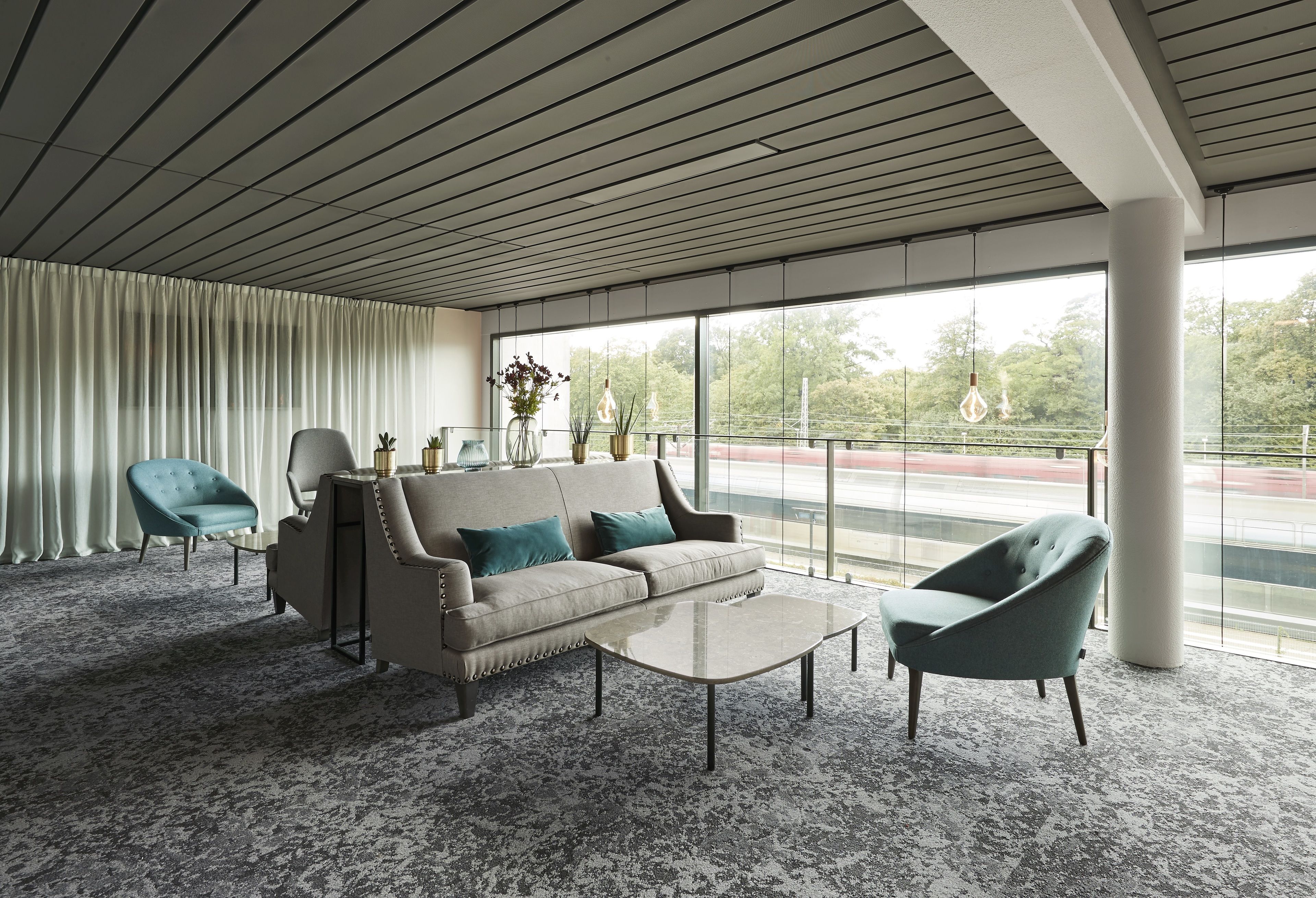 Una sala moderna con un sofá, dos sillones, una mesa de centro y un ventanal con vistas a zonas verdes y una carretera.