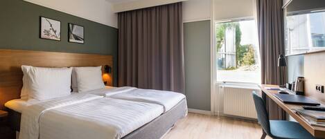 A hotel room with a large bed, a desk with a chair, a window with curtains, and a radiator.