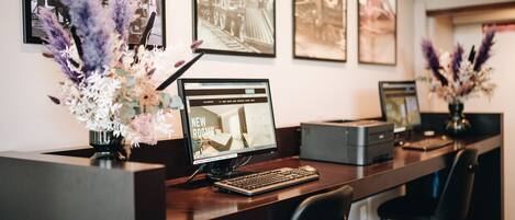A reception desk with a computer monitor displaying 'NEW ROOMS', framed photographs on the wall, and decorative floral arrangements.