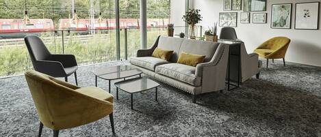 Una sala de estar moderna con un sofá, dos sillones y una mesa de centro. Hay ventanas grandes con vistas a un tren que pasa.