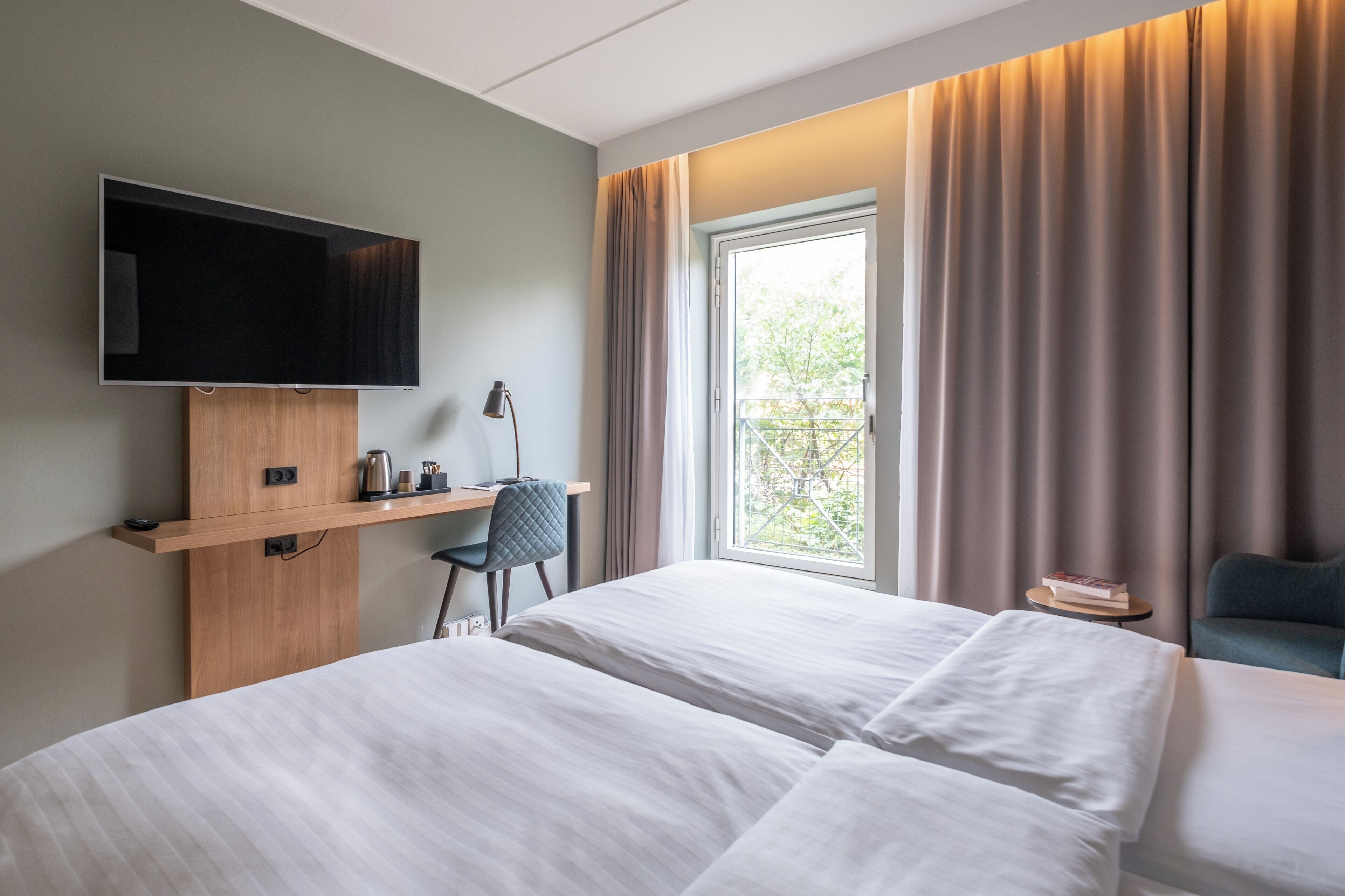 A hotel room with two beds, a TV mounted on a wooden shelf, a desk with a chair, and a window with curtains.