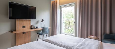 A hotel room with two beds, a TV mounted on a wooden shelf, a desk with a chair, and a window with curtains.