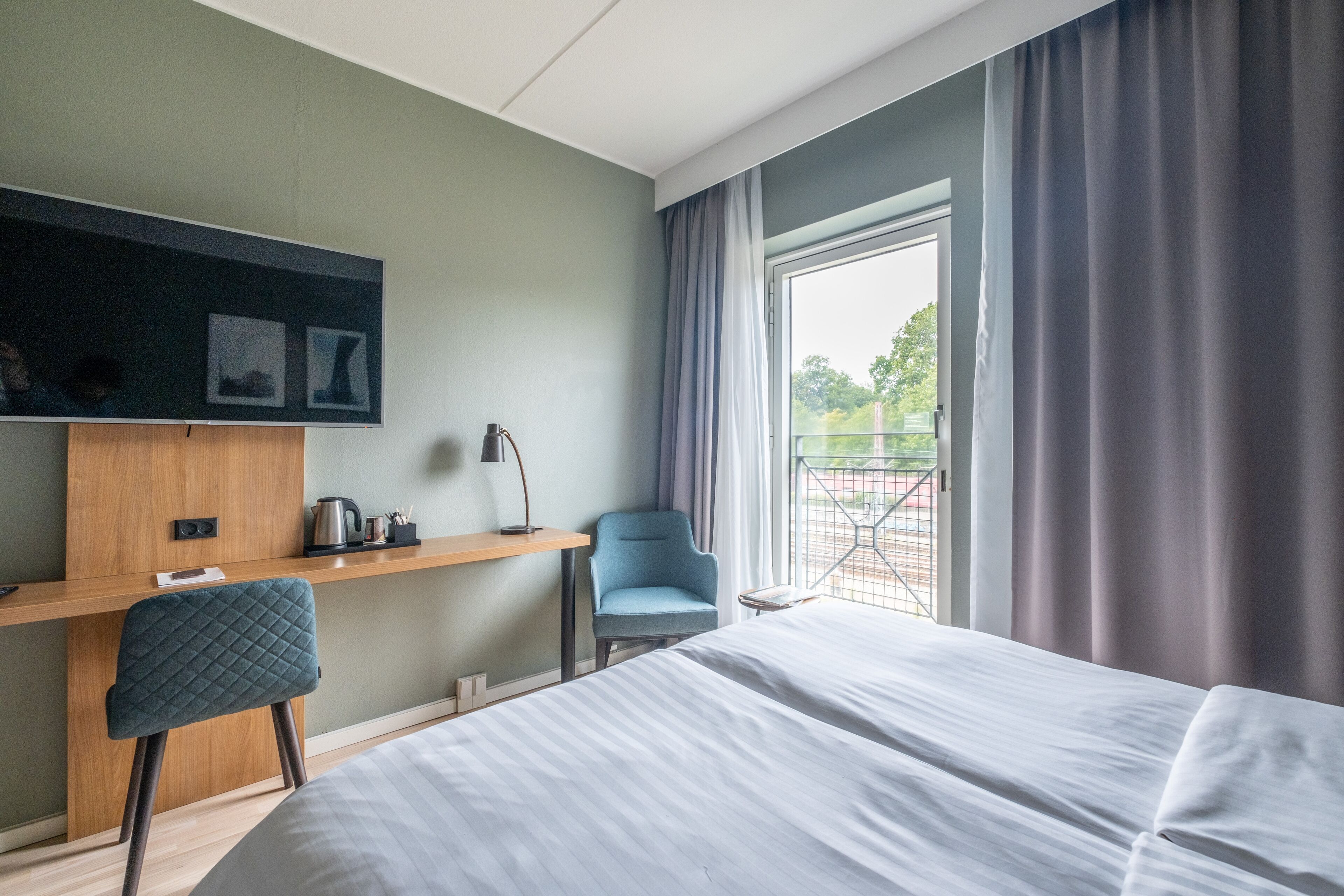 Una habitación de hotel con cama, escritorio con silla, televisor y ventana con cortinas.