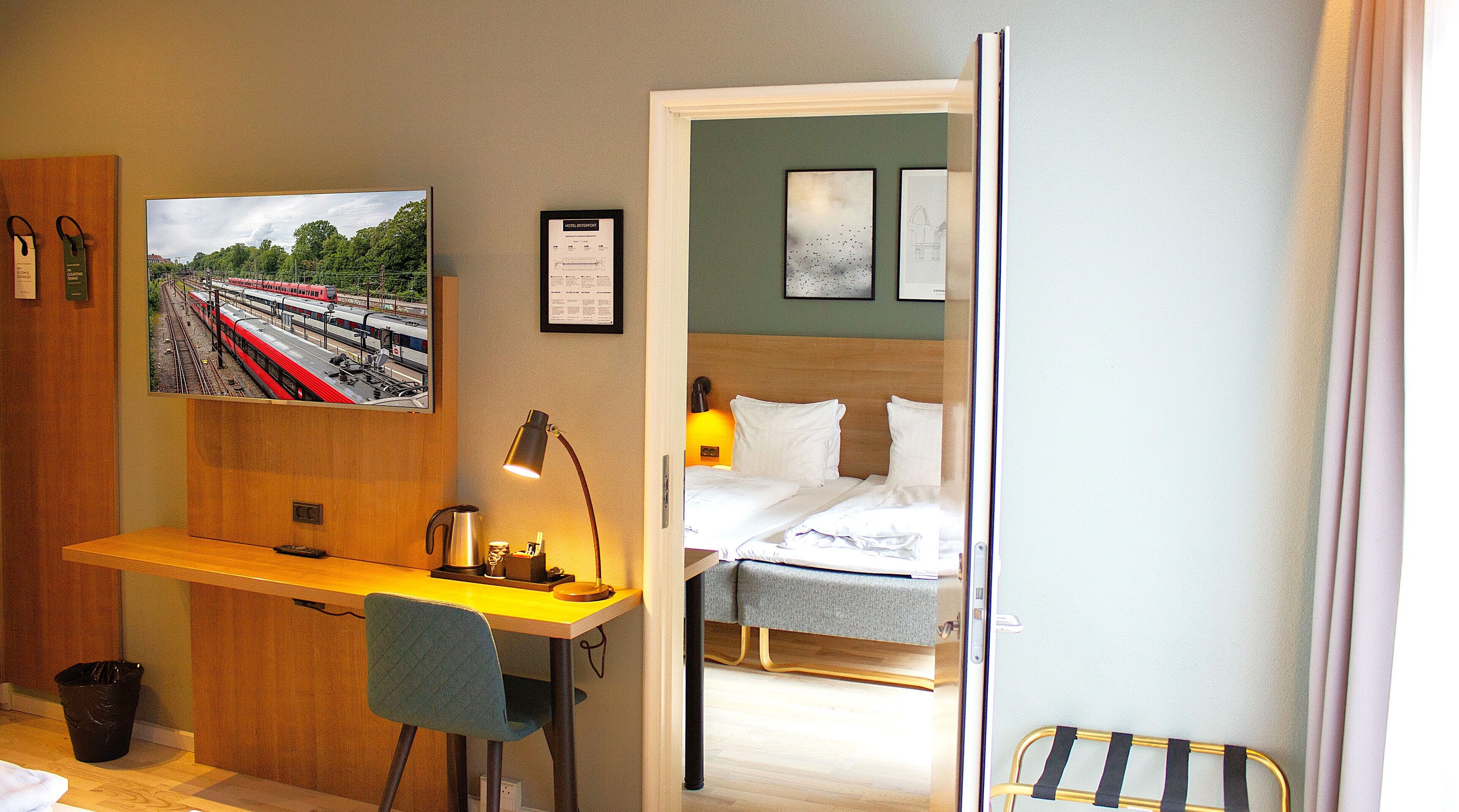 A hotel room with a bed, a desk with a chair, a trash bin, and a wall with a framed picture and a lamp.