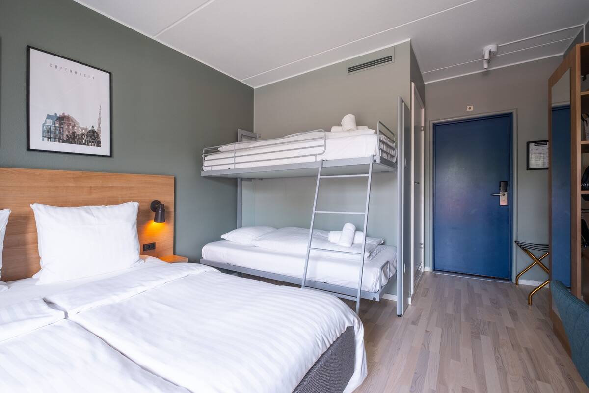 Et hotelværelse med køjeseng, enkeltseng, træsenggavl, et indrammet bybillede, en blå dør og et lille sengebord med en lampe.