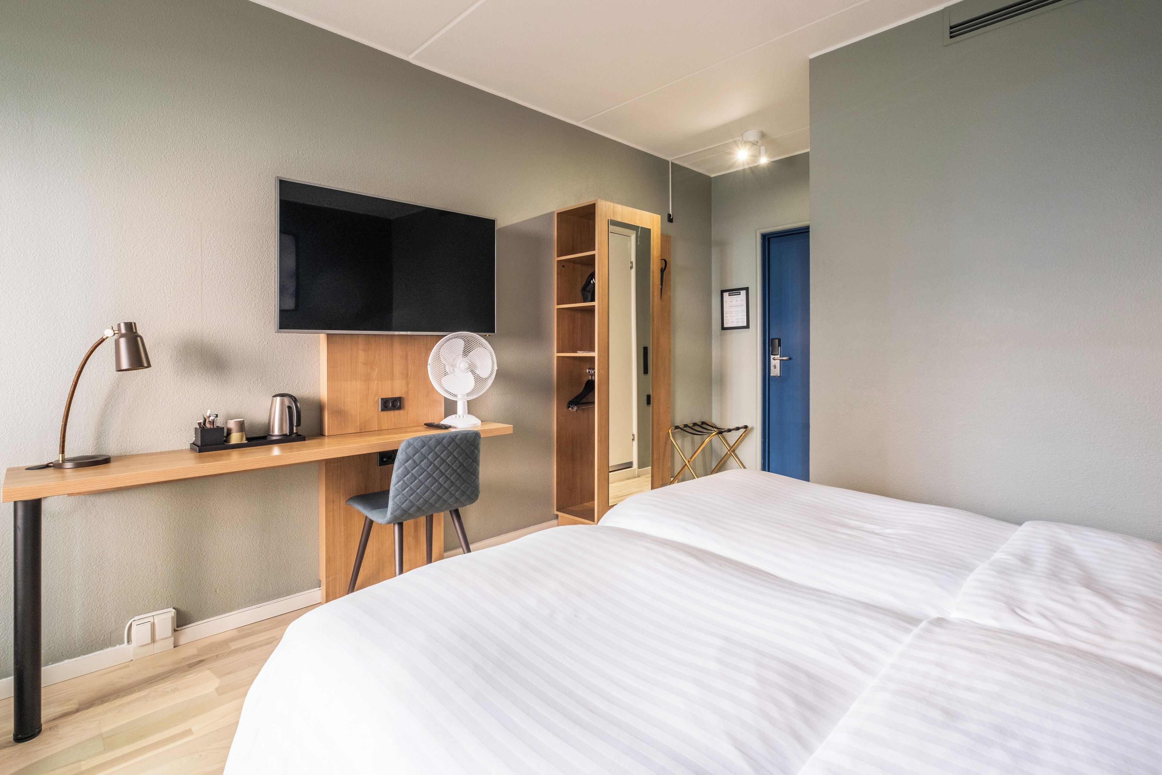 A modern hotel room with a bed, desk, chair, and a wardrobe.