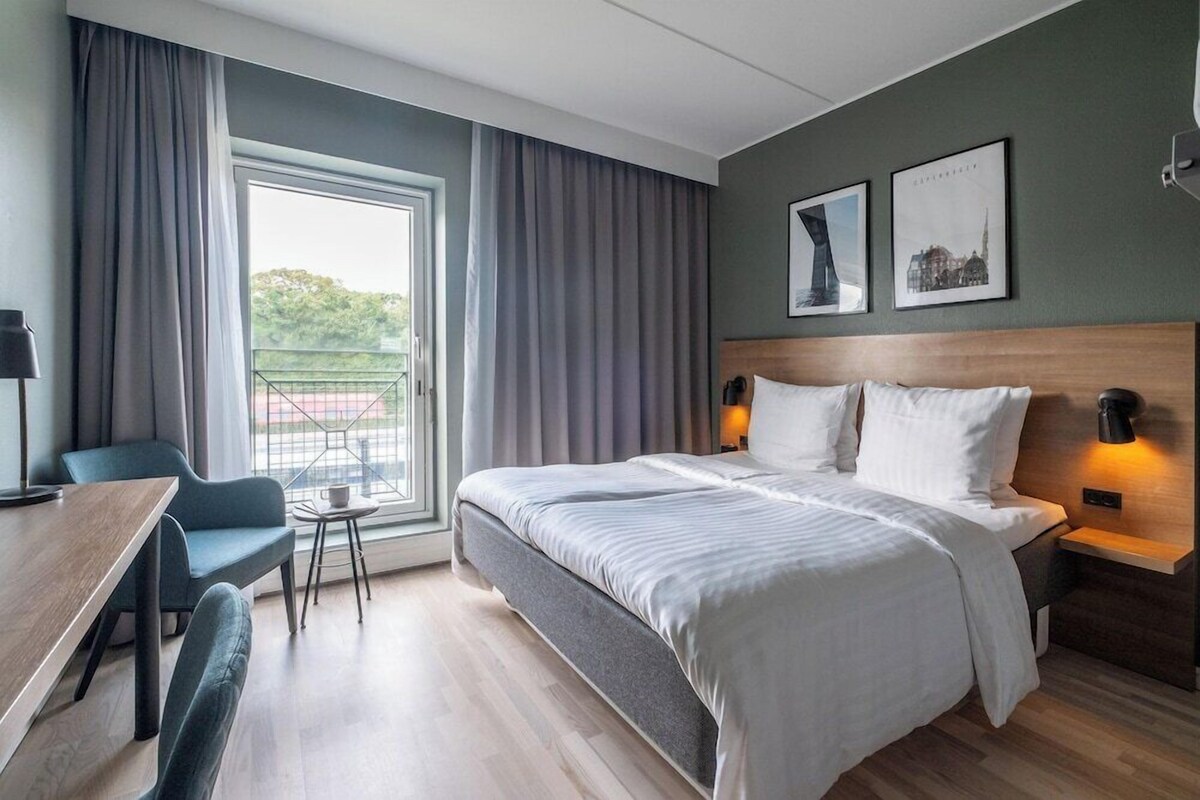A hotel room with a large bed, a desk, a chair, and a balcony with curtains.