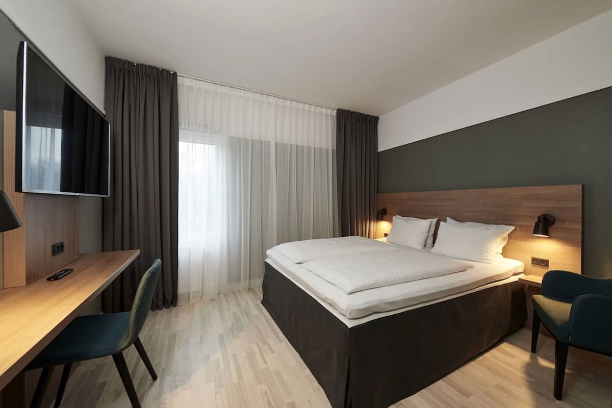 Et moderne hotelværelse med seng, skrivebord, stol, fjernsyn og gardiner.