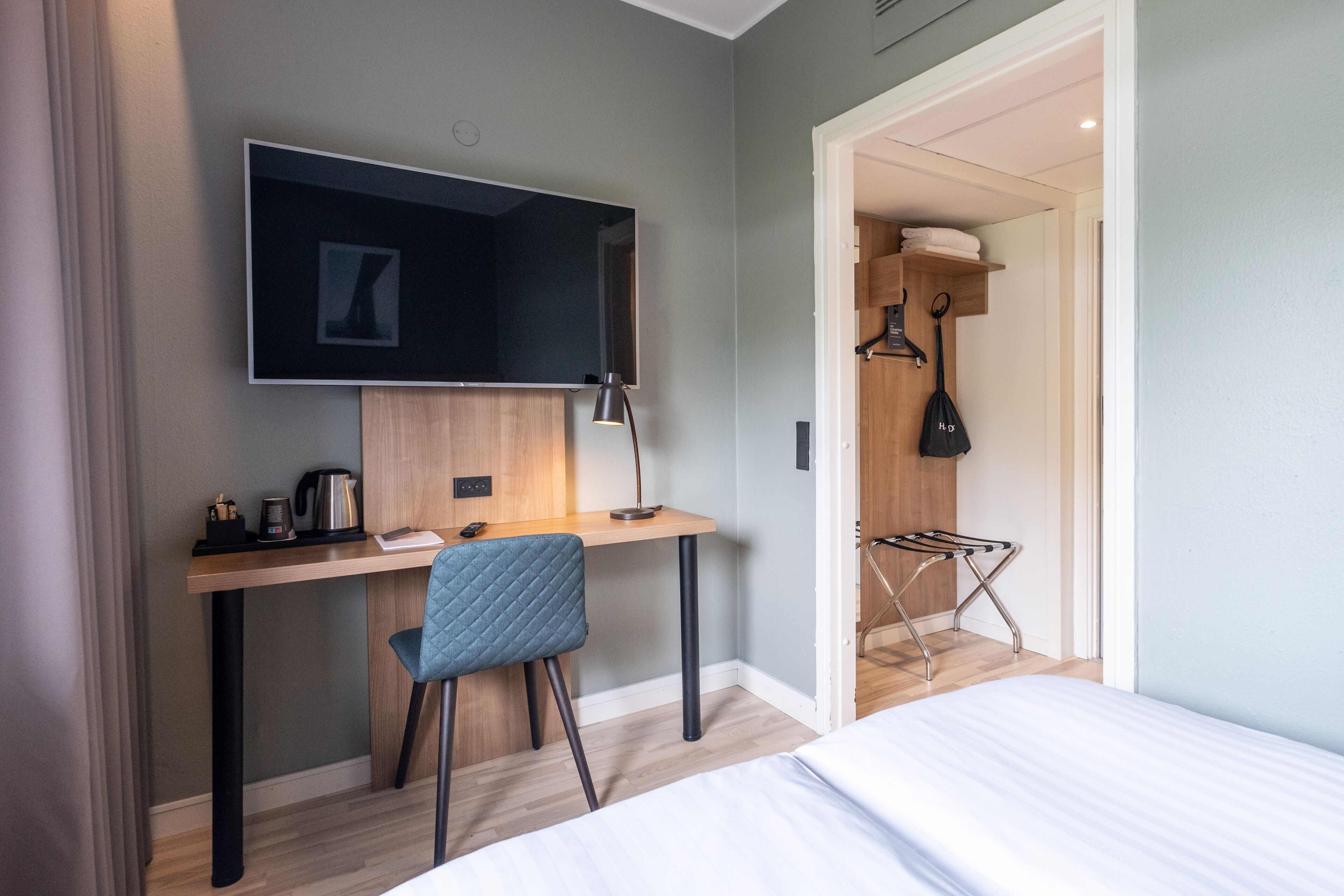 A modern hotel room with a wooden desk, a blue chair, a bed, a TV mounted on the wall, and a walk-in closet with a hanging bag.