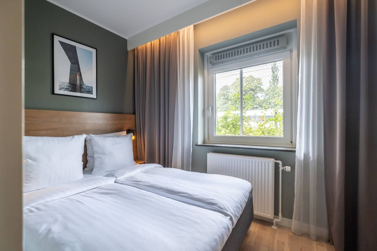 Et hotelværelse med en stor seng, en sengegavl, et vindue med gardiner, en radiator og et billede i ramme på væggen.