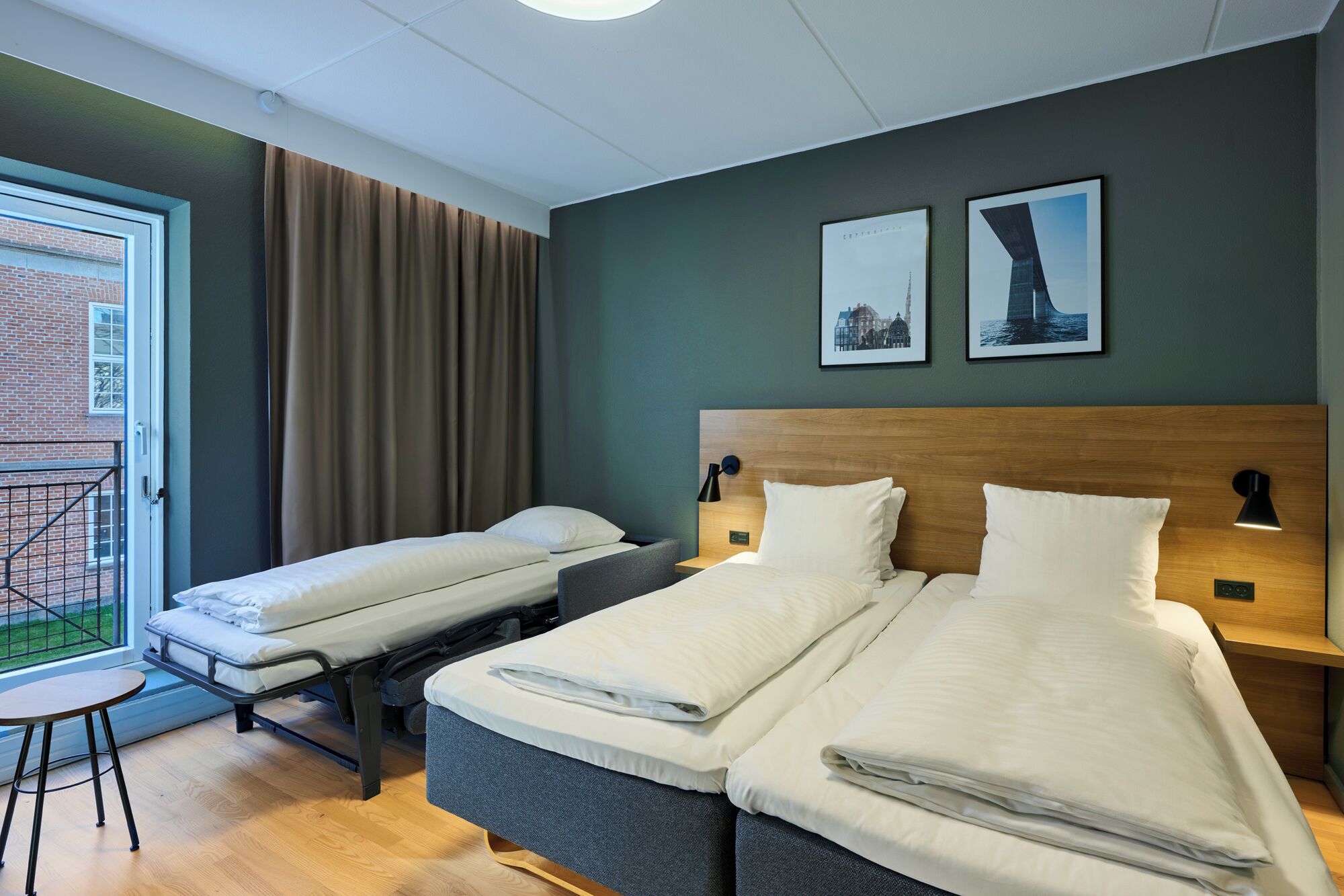 A hotel room with two beds, a small table, a balcony with curtains, and two framed pictures on the wall.