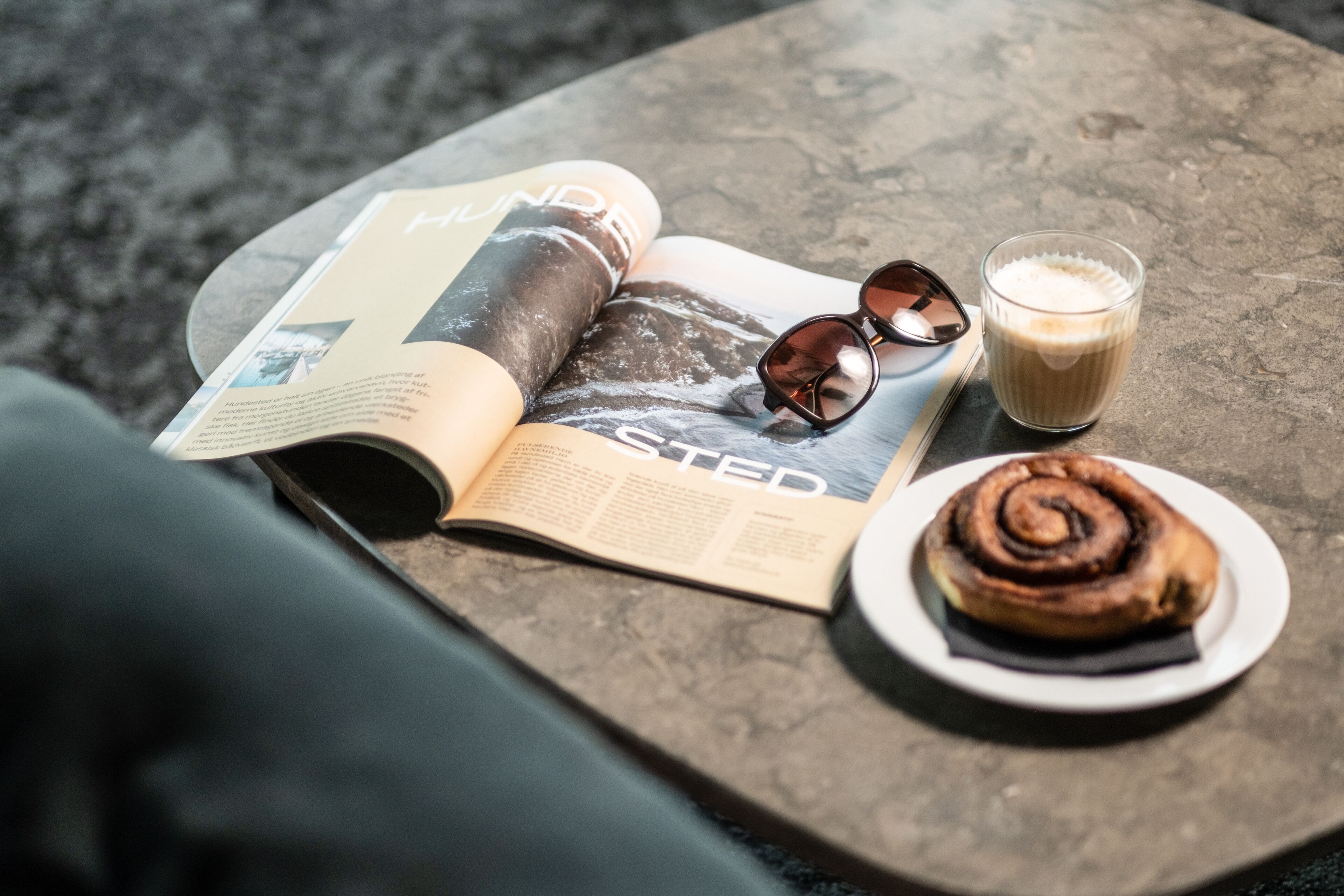 A magazine, a pair of sunglasses, a glass of milk, and a cinnamon roll on a table.