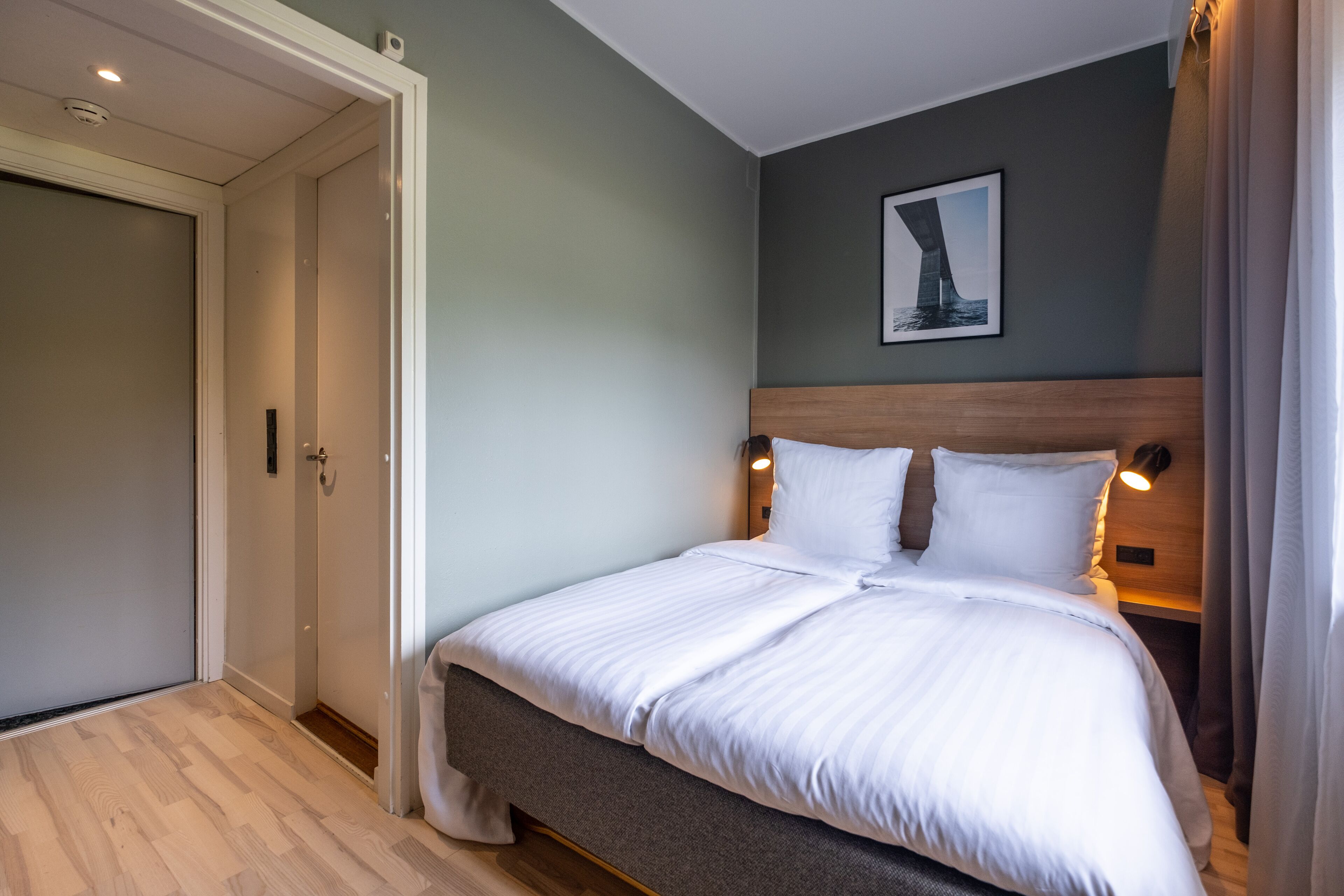 Una cama bien tendida con sábanas blancas en una habitación con paredes verde claro, un cabecero de madera y un cuadro enmarcado en la pared.
