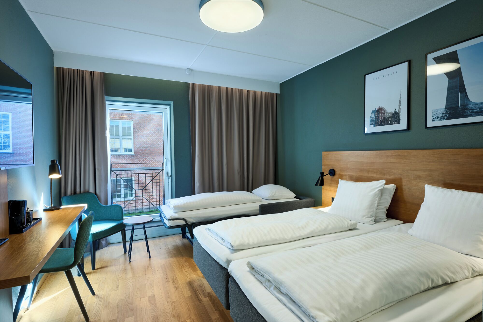 A hotel room with two beds, a desk with a chair, a small table, and a view of a brick building through a window.